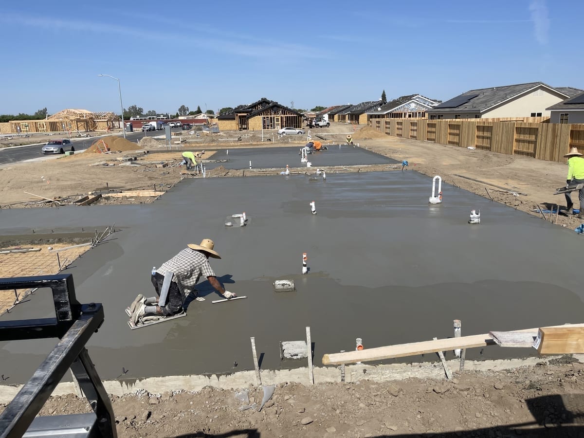 Worker in straw hat hand-troweling a fresh foundation slab pour at a residential development site