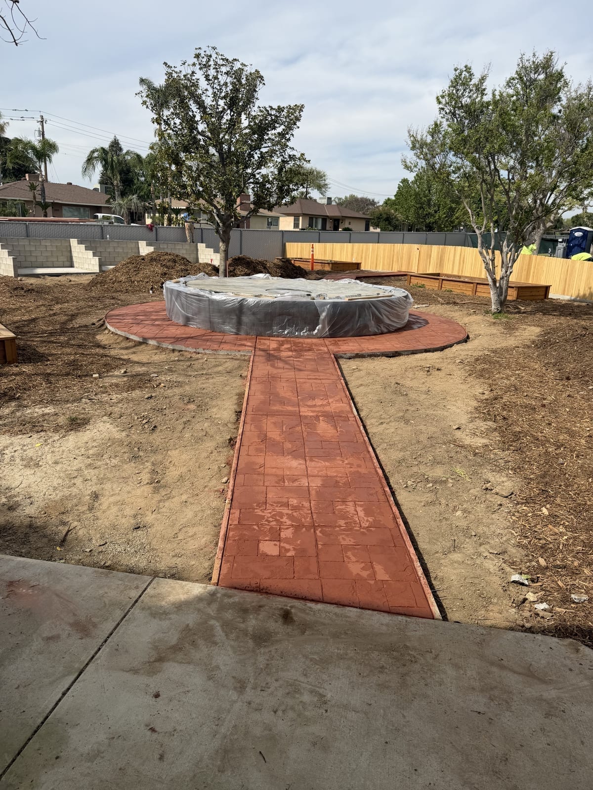 Red stamped concrete walkway leading to a backyard pool installation with mature landscaping